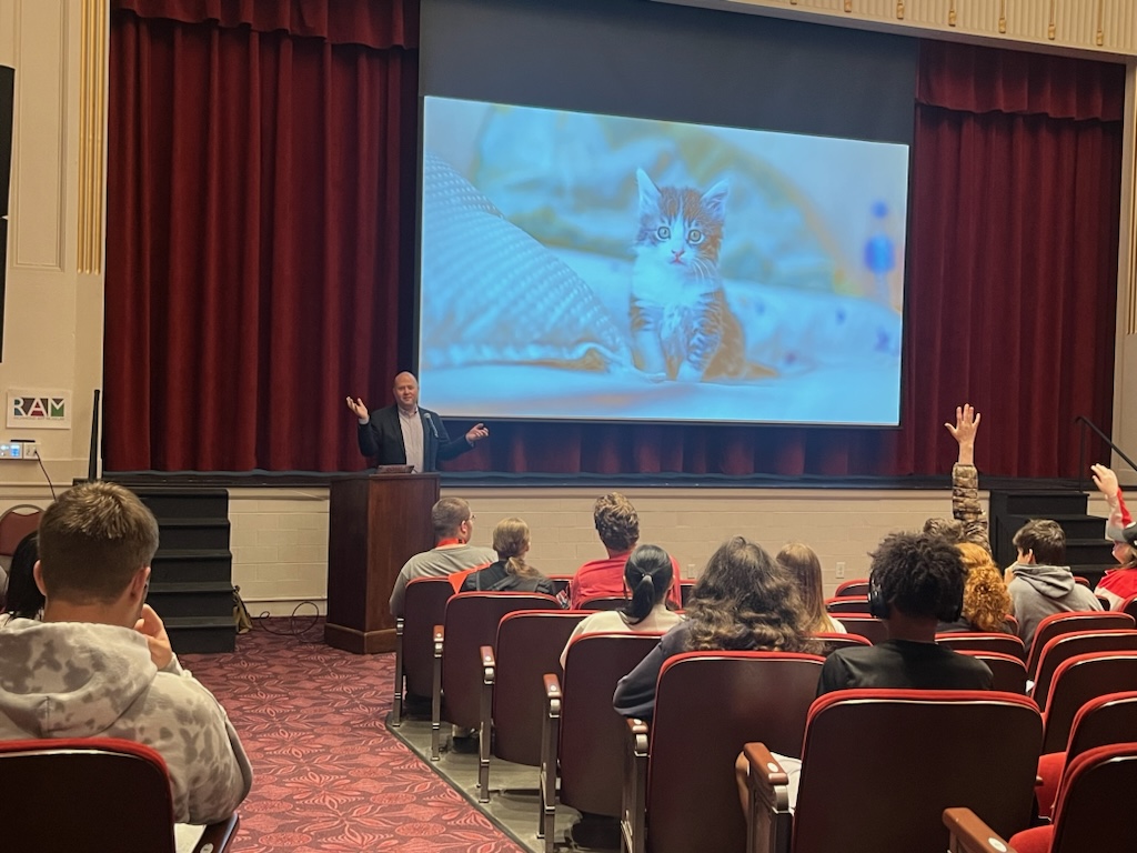 A photo of Chris standing at a podium in front of an auditorium with high school students in the seats, some with hands raised, and on the screen behind him a photo of a kitten sitting on something soft.